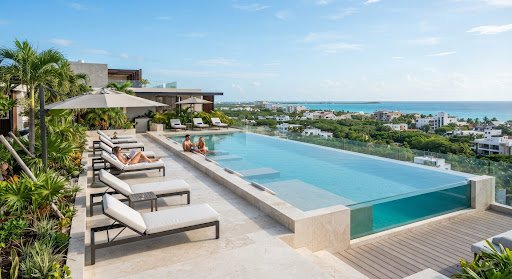 The luxury rooftop pool area of the ANAH Condo building in Playa del Carmen. Features sleek loungers, a pristine infinity pool, and sweeping views of the downtown area. The atmosphere is exclusive, modern, and perfectly captures the high-end tropical vacation vibe.