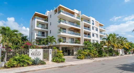 The exterior of Condo Brisas in downtown Playa del Carmen during daytime. A modern, safe residential building with clean white architecture and tropical landscaping. The image conveys security, prime location, and modern comfort under bright, clear Caribbean sunlight.