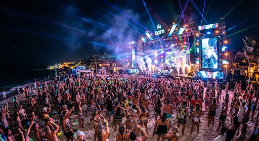 A dynamic, high-energy shot of the BPM Electronic Music Festival on the beach in Playa del Carmen. Crowds of stylish people dancing on the sand under a massive stage setup with colorful lasers and light shows piercing the night sky. The mood is euphoric and vibrant.
